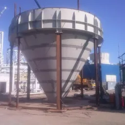 Stożkowy zbiornik przemysłowy, wsparty na stalowych słupach, w trakcie budowy na placu budowy z widocznym dźwigiem i konstrukcjami stalowymi w tle.