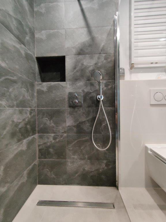 Nowoczesna kabina prysznicowa z szarymi płytkami imitującymi marmur, wbudowaną półką w ścianie, chromowaną deszczownicą i odpływem liniowym w podłodze.