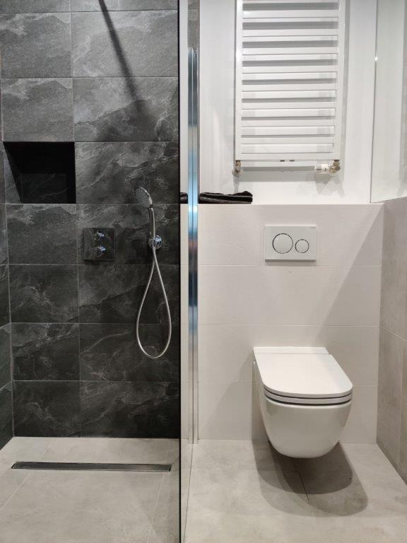 Nowoczesna łazienka z kabiną prysznicową wykończoną grafitowymi płytkami imitującymi marmur, wiszącą toaletą i grzejnikiem drabinkowym, widoczny odpływ liniowy w podłodze.