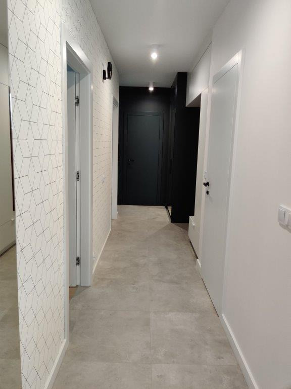 Nowoczesny korytarz z szarą podłogą, białą ścianą z geometrycznym wzorem i ciemnymi drzwiami w głębi.