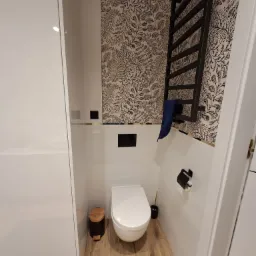 Nowoczesna toaleta z białymi płytkami, wzorzystą tapetą w liście, czarnym grzejnikiem drabinkowym z niebieskim ręcznikiem, drewnopodobną podłogą i czarnymi akcentami.