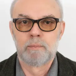 Portret starszego mężczyzny z siwą brodą i włosami, w okularach z czarnymi oprawkami, ubranego w szarą koszulę i tweedową marynarkę, z przypinką orła na klapie.