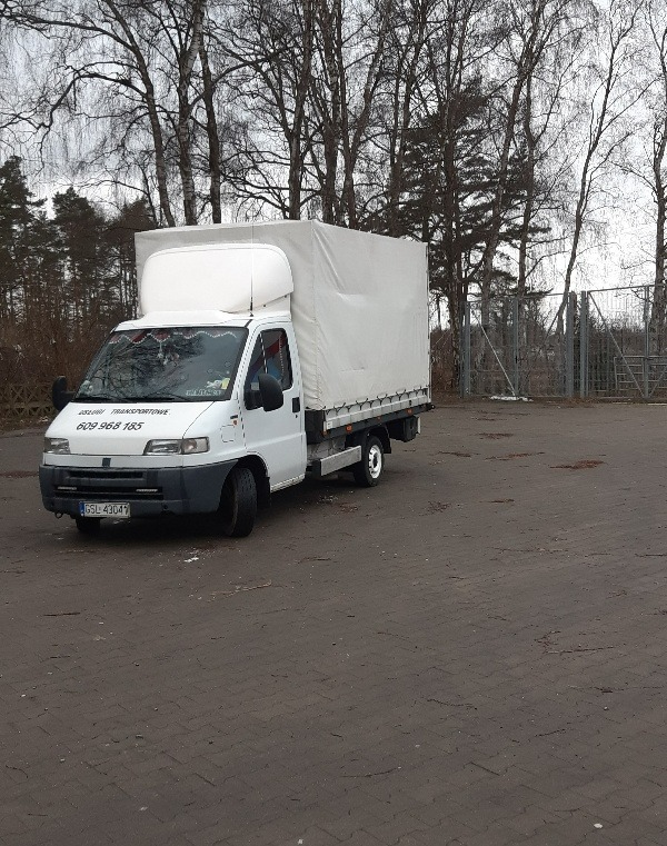 Biały samochód dostawczy z plandeką na parkingu, widoczne drzewa w tle, numer telefonu na przedniej szybie.