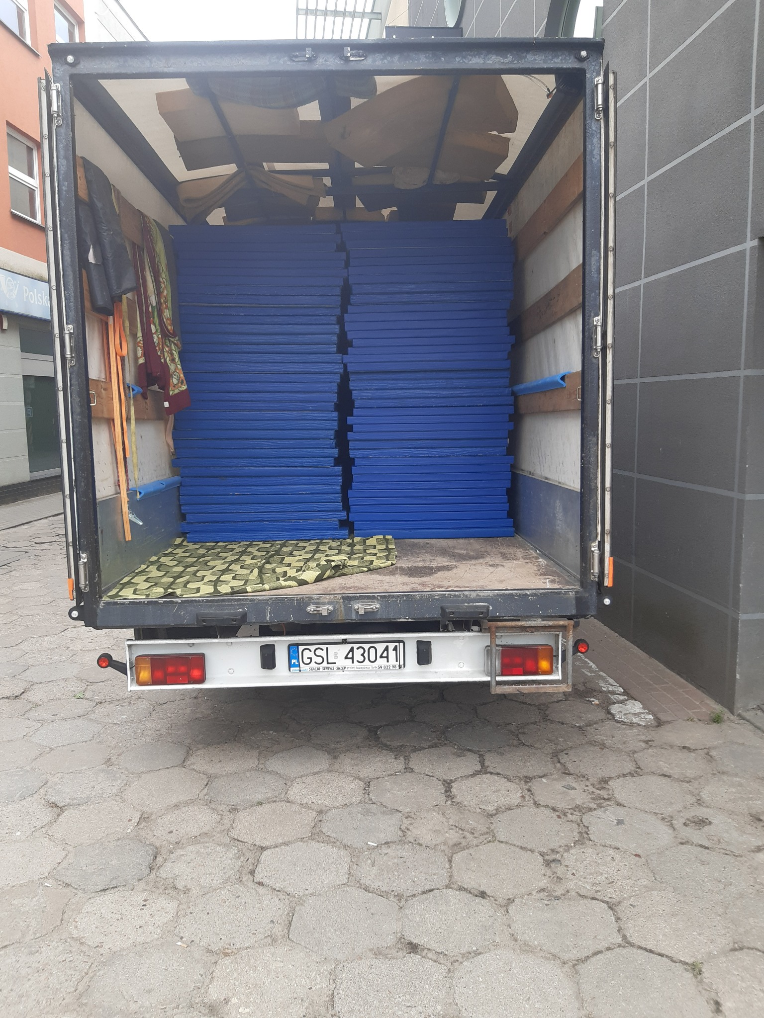 Wnętrze busa transportowego wypełnione stosem niebieskich mat, widok z tyłu na otwarte drzwi i tablicę rejestracyjną GSL 43041, z elementami wyposażenia busa po bokach.