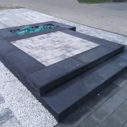 Nowoczesny taras z kostki brukowej w odcieniach szarości, z elementami dekoracyjnymi z białego żwiru i szkła w wyznaczonym obszarze, tworzący wielopoziomową przestrzeń.