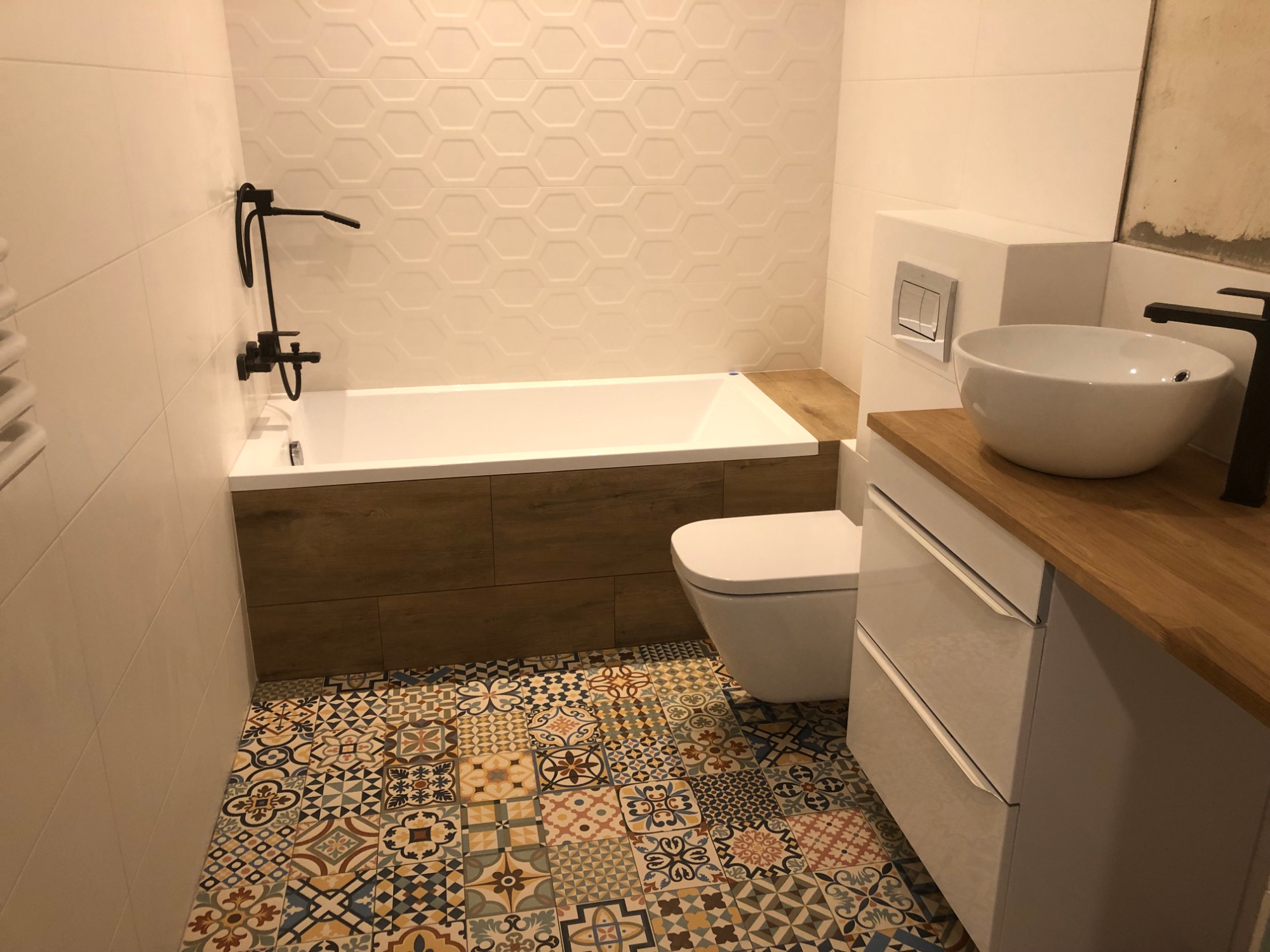 Nowoczesna łazienka z wanną obudowaną drewnem, białą ceramiką, szafką z drewnianym blatem i wzorzystą, kolorową podłogą.