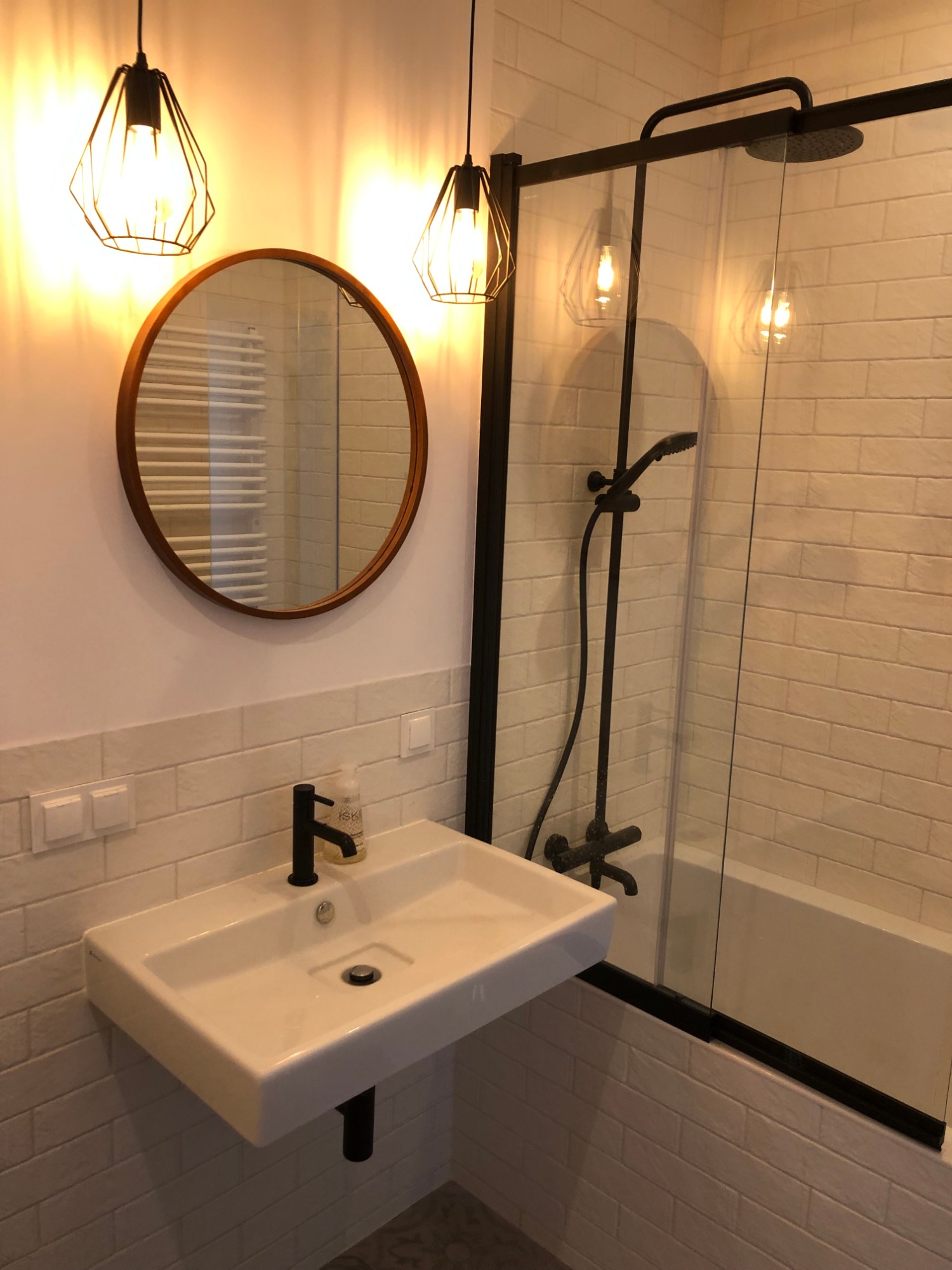 Nowoczesna łazienka z białymi cegiełkowymi płytkami, okrągłym lustrem w drewnianej ramie, wiszącą umywalką z czarną armaturą i kabiną prysznicową typu walk-in z czarnymi profilami.