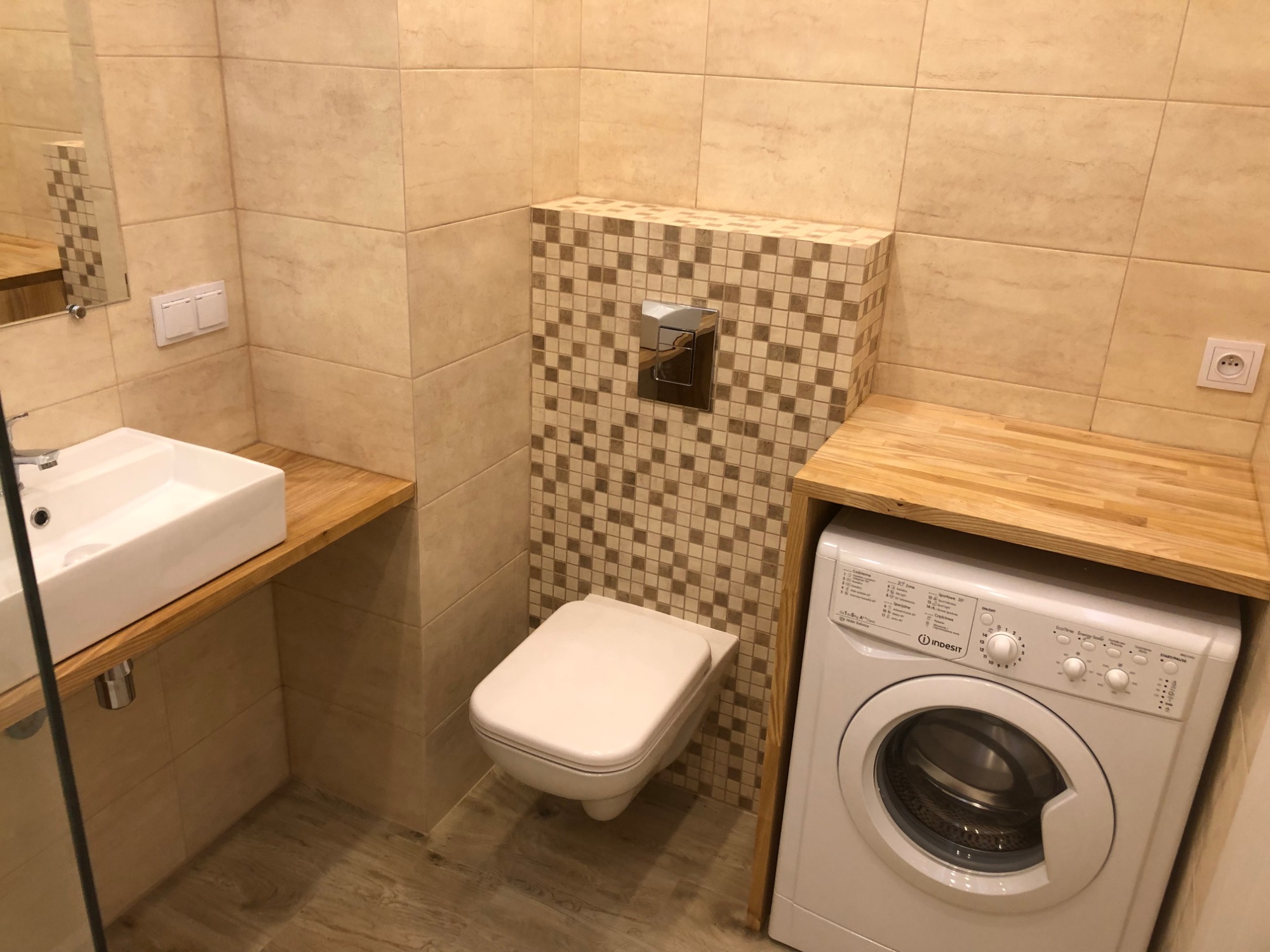Jasna łazienka z beżowymi płytkami, wiszącą toaletą obudowaną mozaiką, białą umywalką nablatową na drewnianym blacie i pralką Indesit pod blatem.