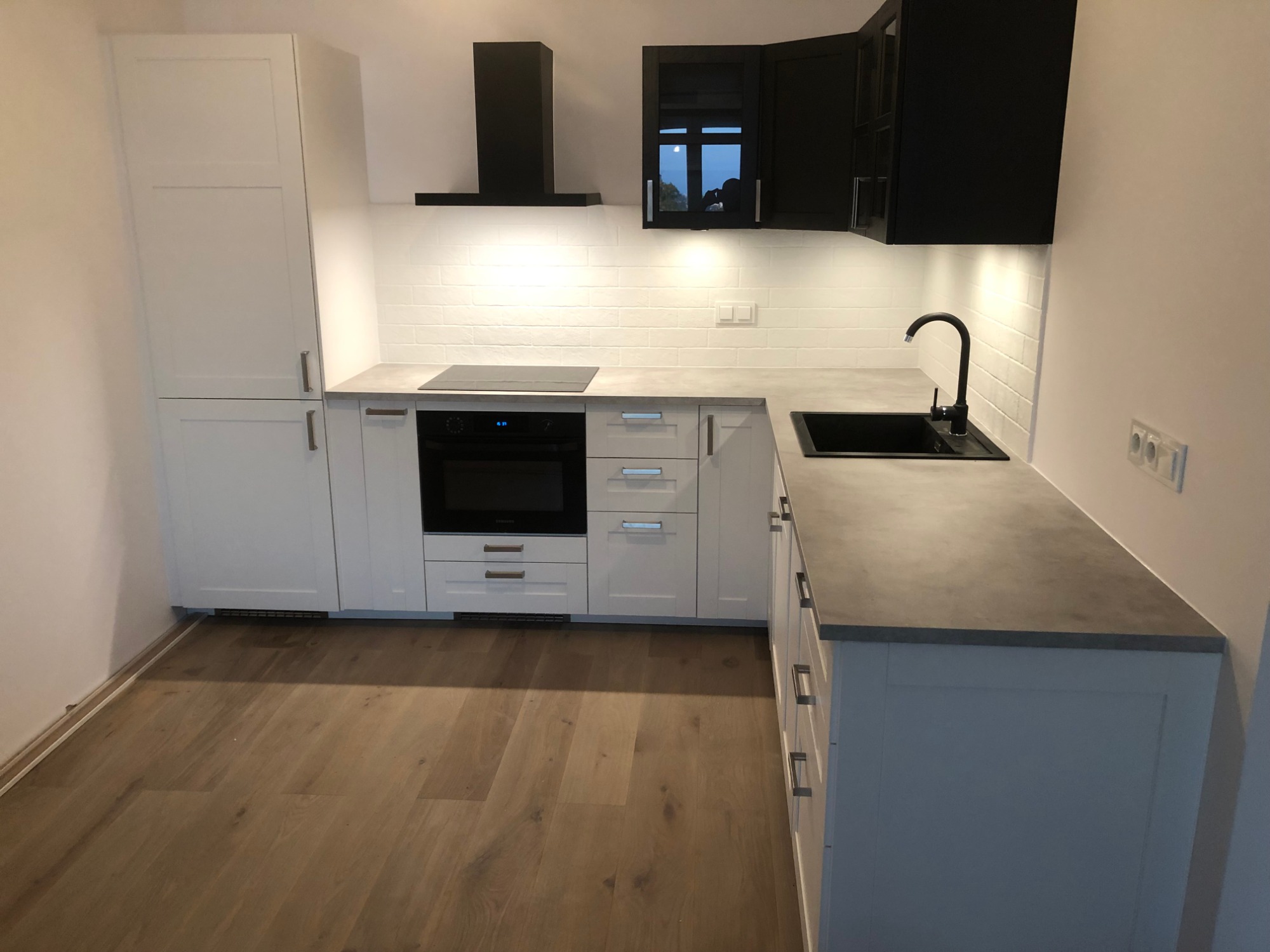 Nowoczesna kuchnia w bieli i czerni: białe szafki dolne i wysoka zabudowa, czarne szafki wiszące i okap, szary blat, białe płytki na ścianie i drewniana podłoga.