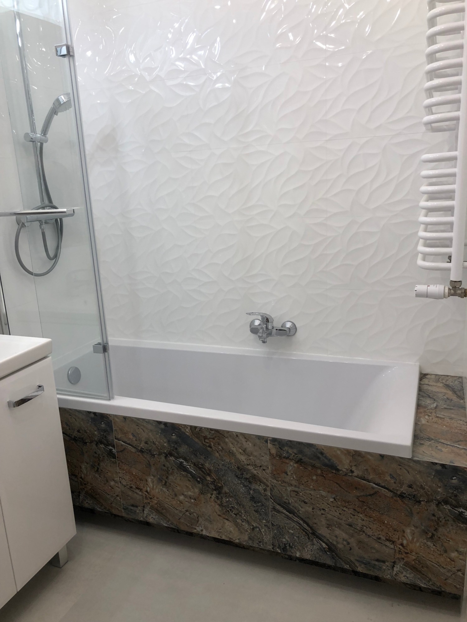 Biała wanna z szklanym parawanem i chromowaną armaturą na tle ściany z białych płytek o falistej strukturze; dolna część obudowana płytkami imitującymi marmur.