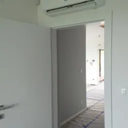 Biały klimatyzator Gree LOMO Luxury zamontowany na ścianie nad drzwiami w jasnym, minimalistycznym wnętrzu, z folią ochronną na podłodze.