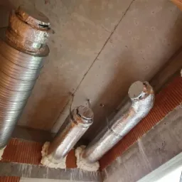 Montaż srebrnych, elastycznych przewodów wentylacyjnych w surowym stanie budynku z cegły i betonu, zabezpieczonych pianką montażową.