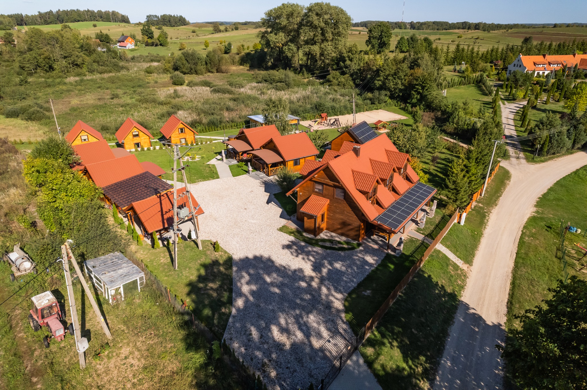 Widok z drona na kompleks drewnianych domków z czerwonymi dachami i panelami słonecznymi, otoczonych zielenią i polami.