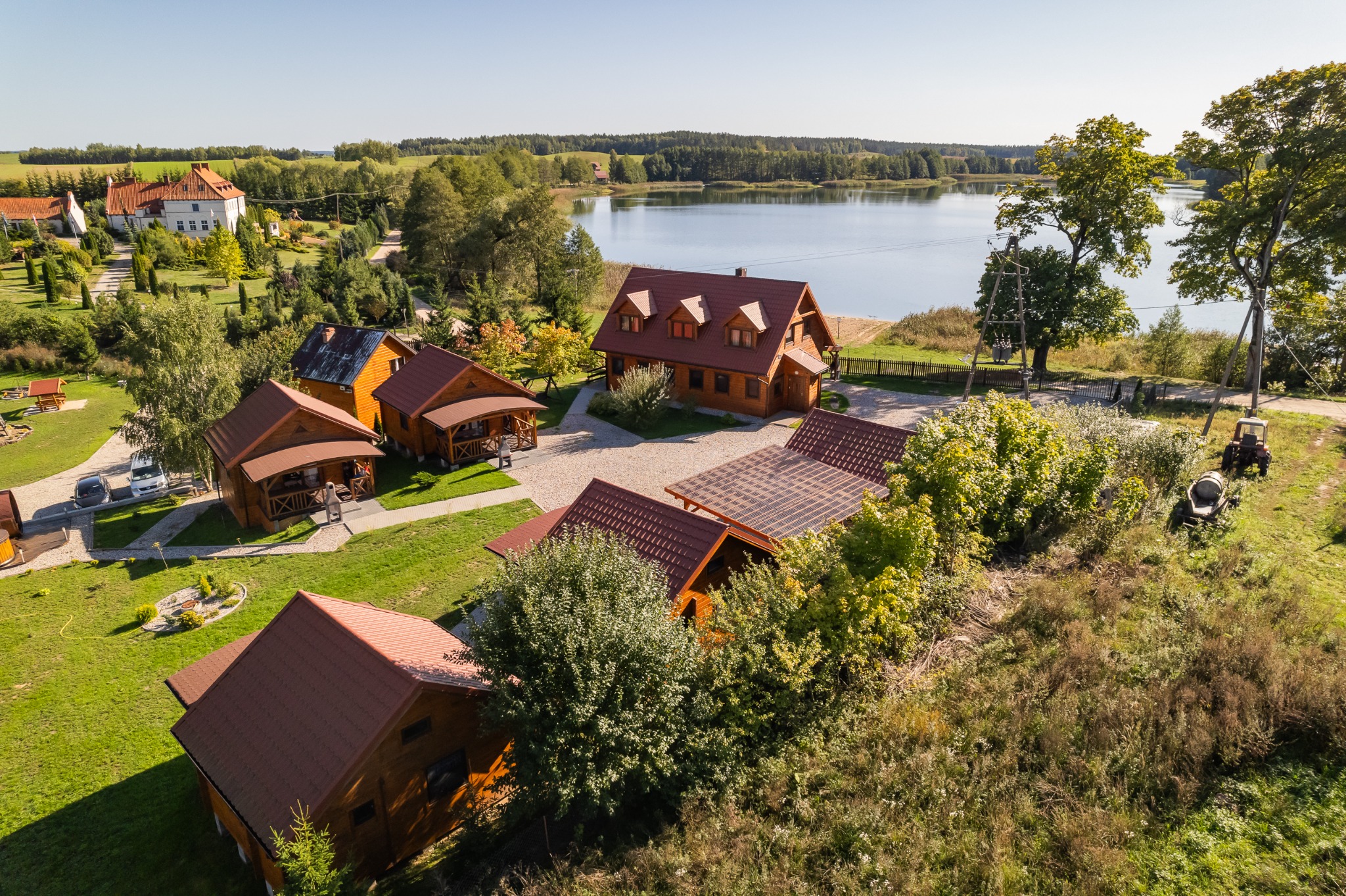 Panoramiczne ujęcie z drona na kompleks drewnianych domków letniskowych z czerwonymi dachami, położonych nad jeziorem, z widoczną linią brzegową i lasem w tle.