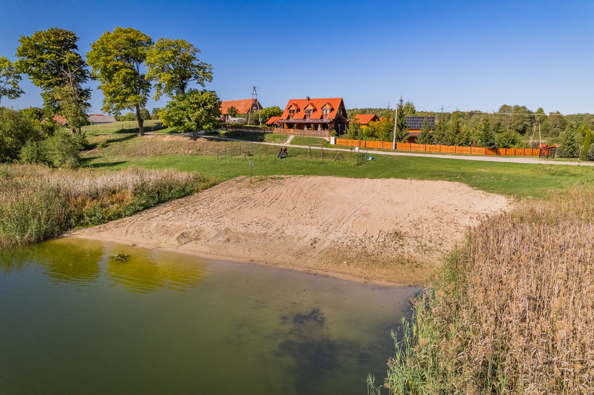 Widok z lotu ptaka na drewniany dom z czerwonym dachem, plażę i staw w słoneczny dzień.