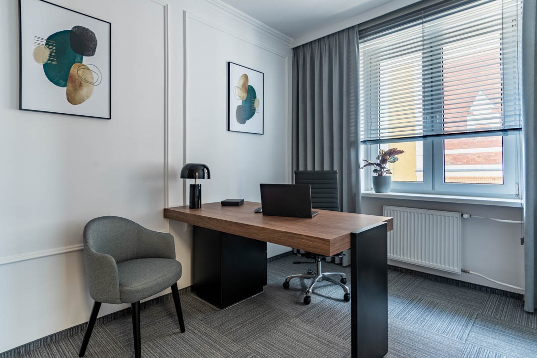Biuro z drewnianym biurkiem, czarną lampką, szarym krzesłem i abstrakcyjnymi obrazami na ścianie, oświetlone światłem z okna z żaluzjami.