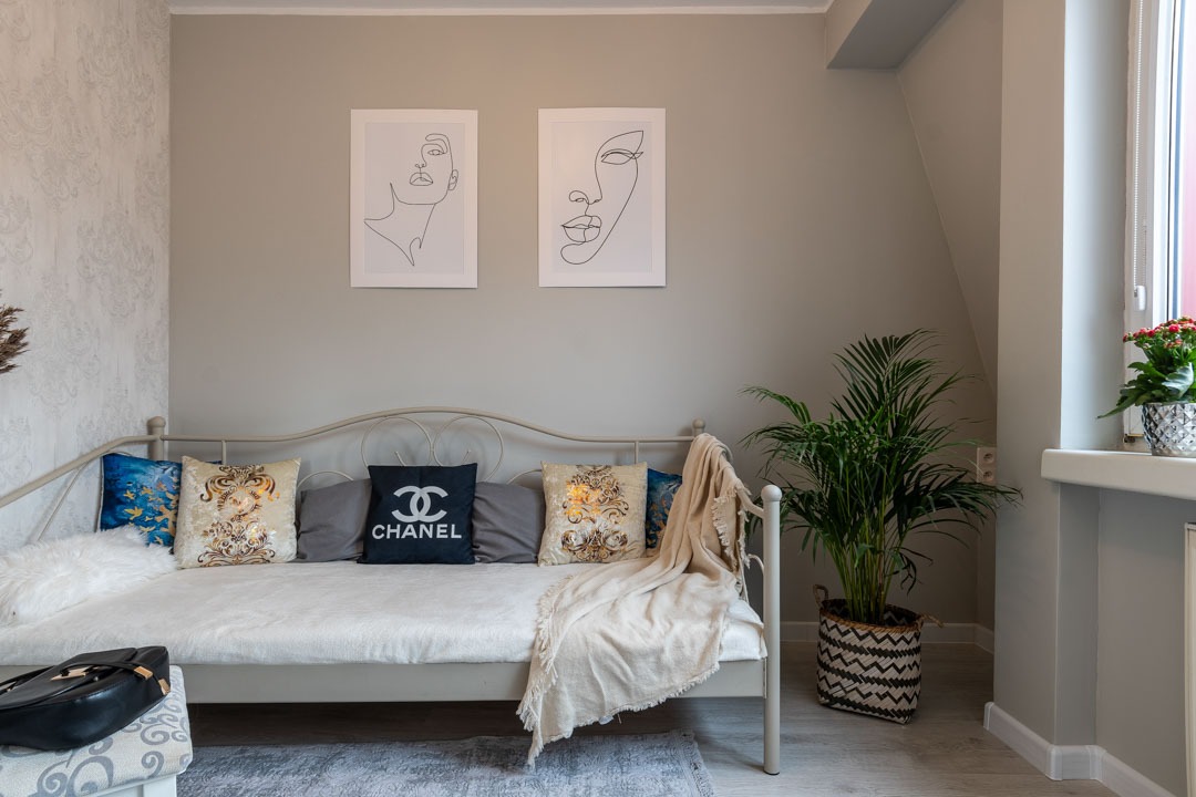 Jasnoszary pokój z metalową sofą, poduszkami dekoracyjnymi (w tym jedna z logo Chanel) i dwoma minimalistycznymi portretami na ścianie, obok okna stoi doniczka z kwiatami, a obok sofy palma...