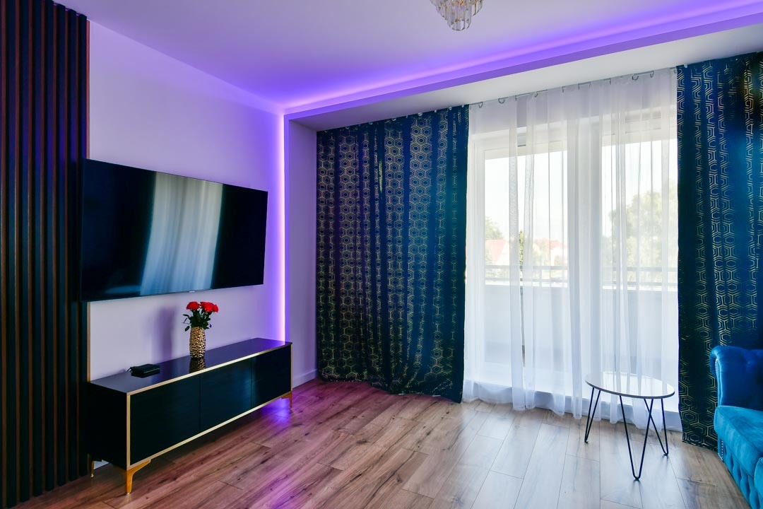 Salon z drewnianą podłogą, czarną szafką RTV z bukietem czerwonych róż, telewizorem, zasłonami w geometryczne wzory i oświetleniem LED w kolorze fioletowym.