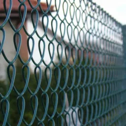 Zielona siatka ogrodzeniowa z bliska, widoczny dom w tle. Perspektywa z dołu ukazuje regularny wzór oczek i solidną konstrukcję. Dobre oświetlenie.