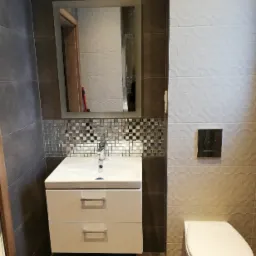 Nowoczesna łazienka z białym zlewem z szafką, lustrem z podświetleniem, mozaiką nad zlewem i podwieszanym WC na tle szaro-białych płytek.