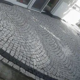 Granitowa nawierzchnia ułożona w łukowy wzór, obrzeża z ciemnej kostki. Widok na budynek z białymi drzwiami i fragment ogrodu z ławką.