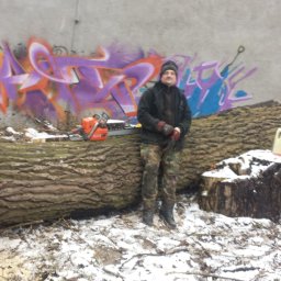 ANKER-GROUP Piotr KOTWICA - Mężczyzna w zimowym ubraniu stoi obok pnia drzewa z piłą łańcuchową, na tle ściany z graffiti, pozostałości po wycince drzewa w zimowej scenerii.