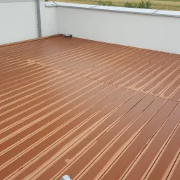 Kompozytowa deska tarasowa w odcieniach brązu, ułożona na balkonie z widocznym odpływem wody deszczowej i fragmentem białej ściany z metalowym orynnowaniem.