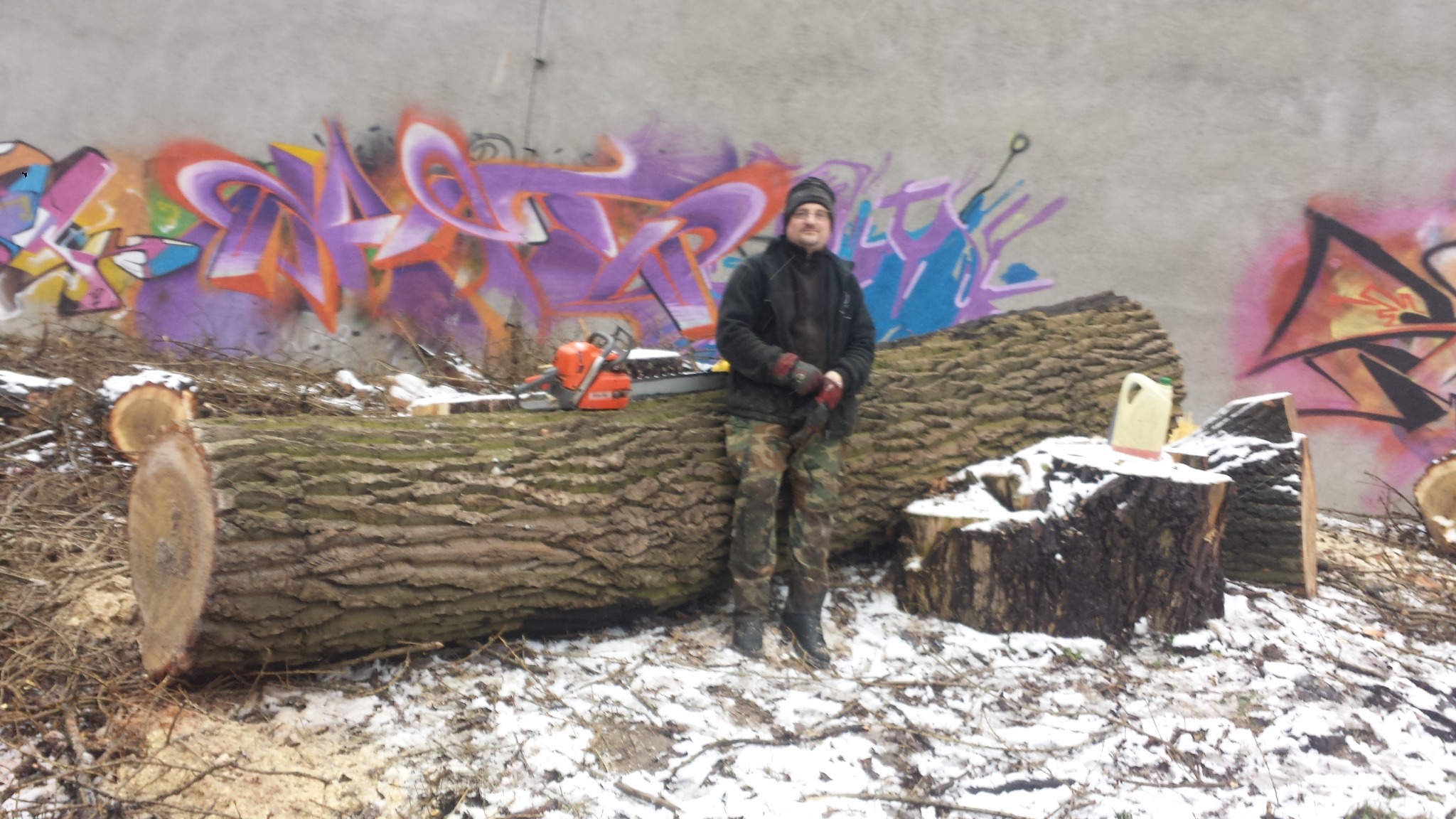 Mężczyzna w zimowym ubraniu stoi obok pnia drzewa z piłą łańcuchową, na tle ściany z graffiti, pozostałości po wycince drzewa w zimowej scenerii.