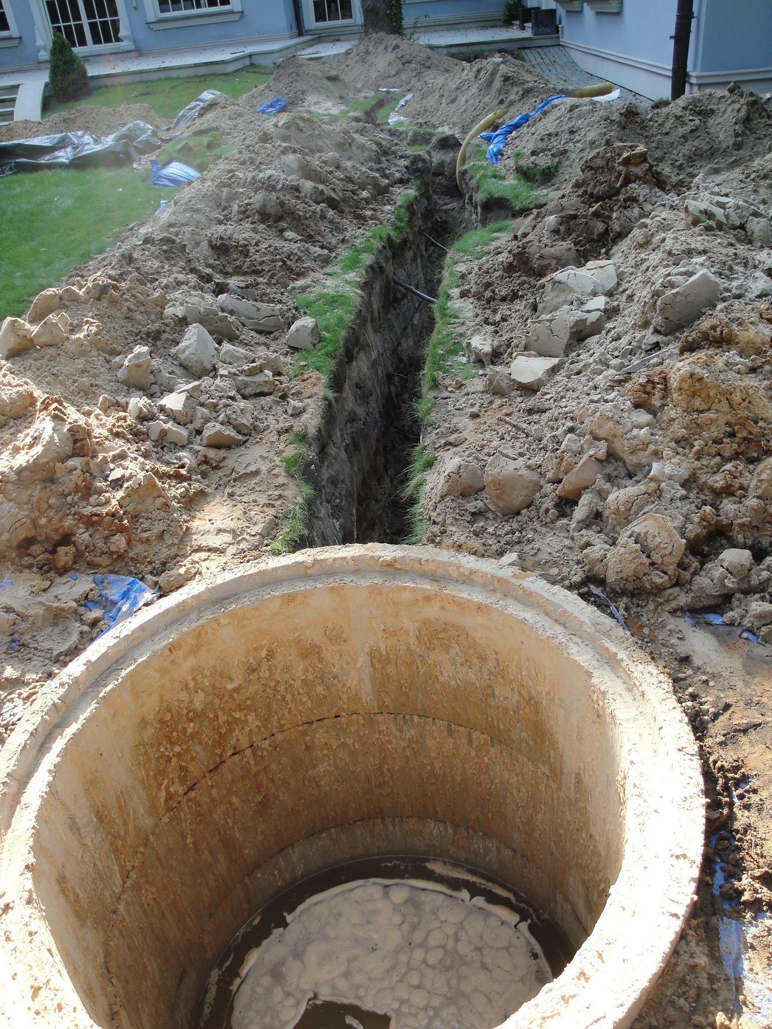 Betonowy krąg studni kanalizacyjnej na pierwszym planie, w tle wykopany wąski rów w ziemi, fragment trawnika i domu w tle.