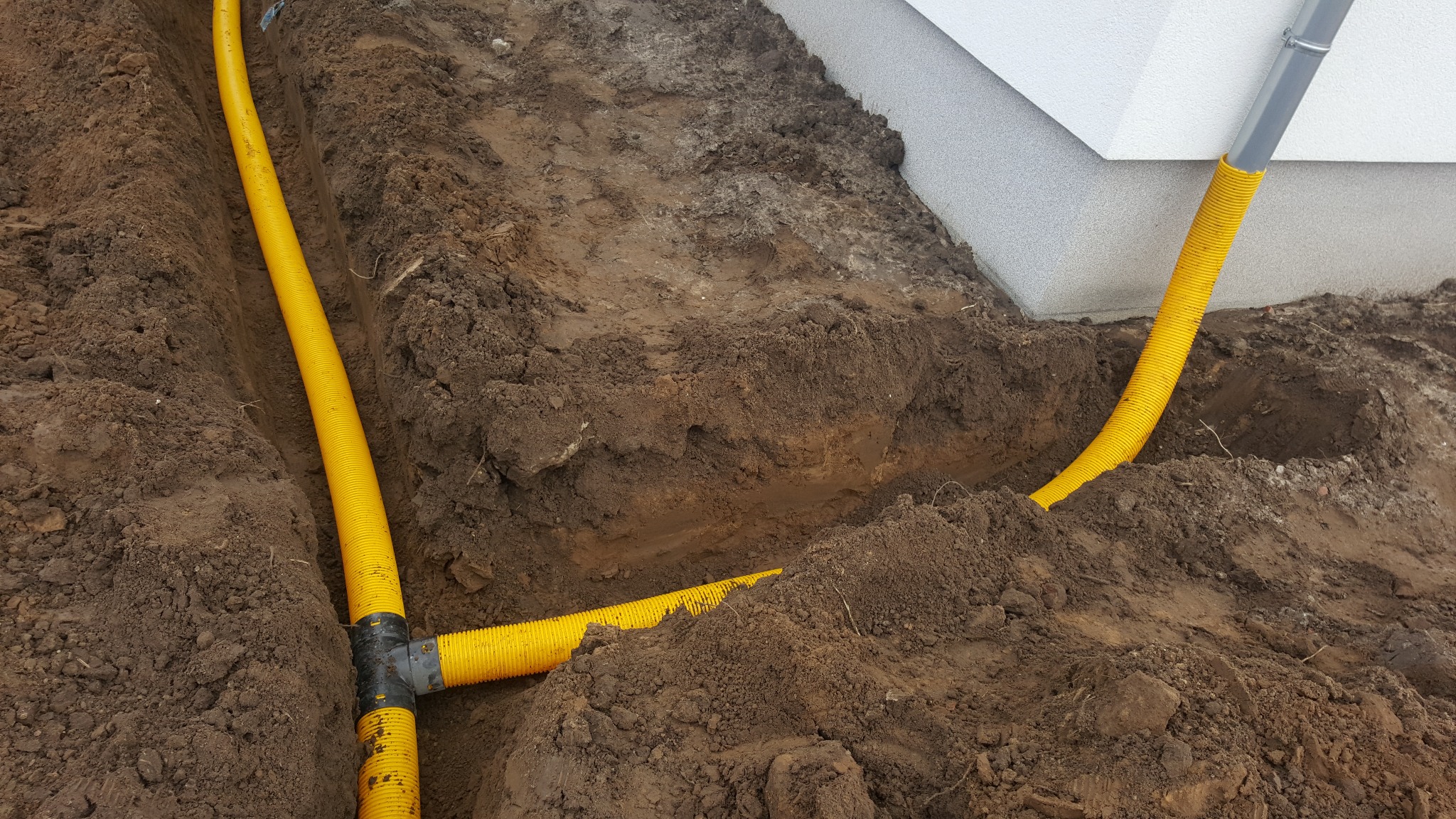 Żółta rura drenażowa ułożona w wykopie obok fundamentu budynku, widoczne złącza i fragment rury spustowej.