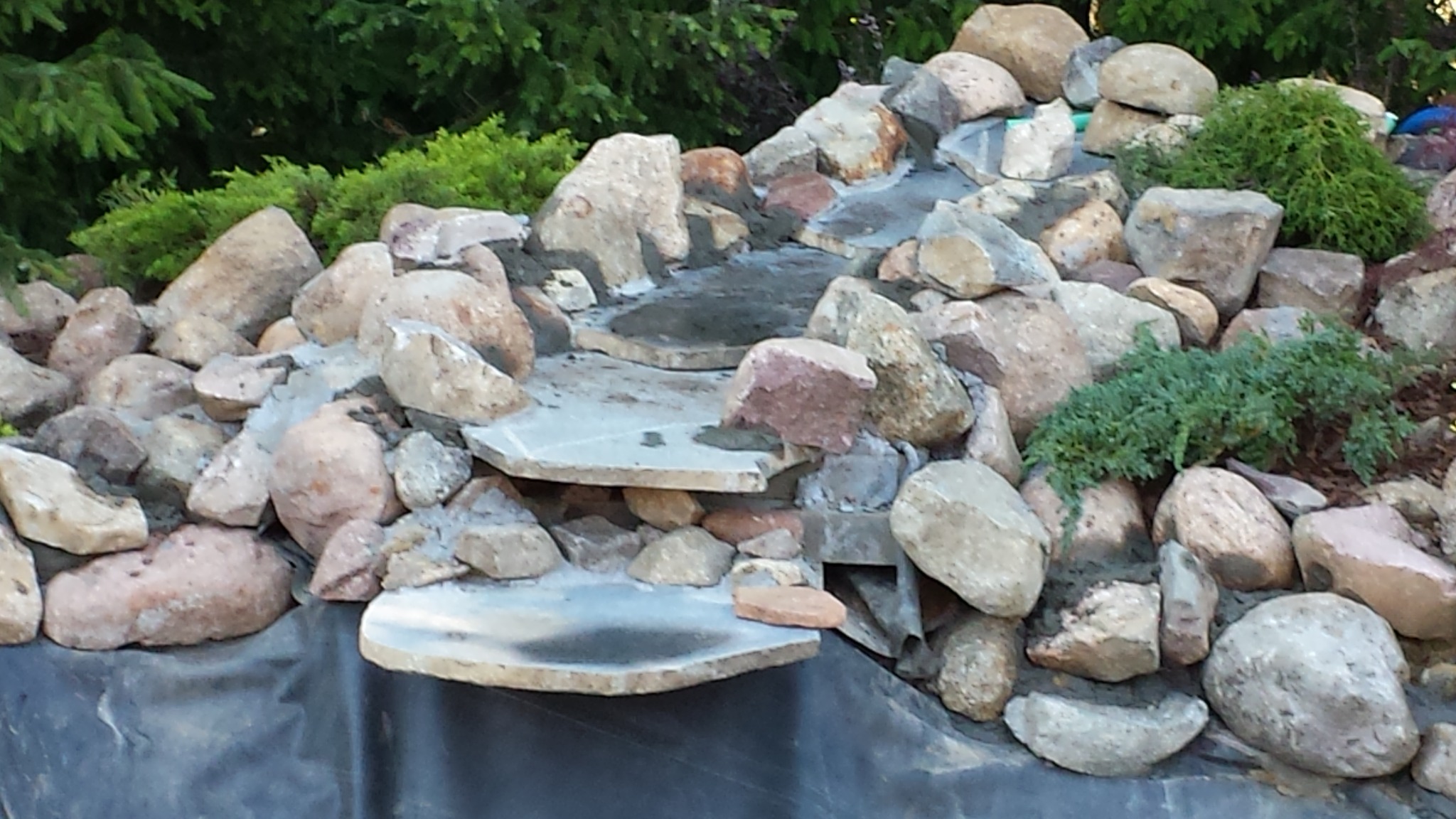 Kaskada wodna w ogrodzie w trakcie budowy, z użyciem kamieni różnej wielkości i płyt kamiennych, widoczna folia hydroizolacyjna i roślinność w tle.