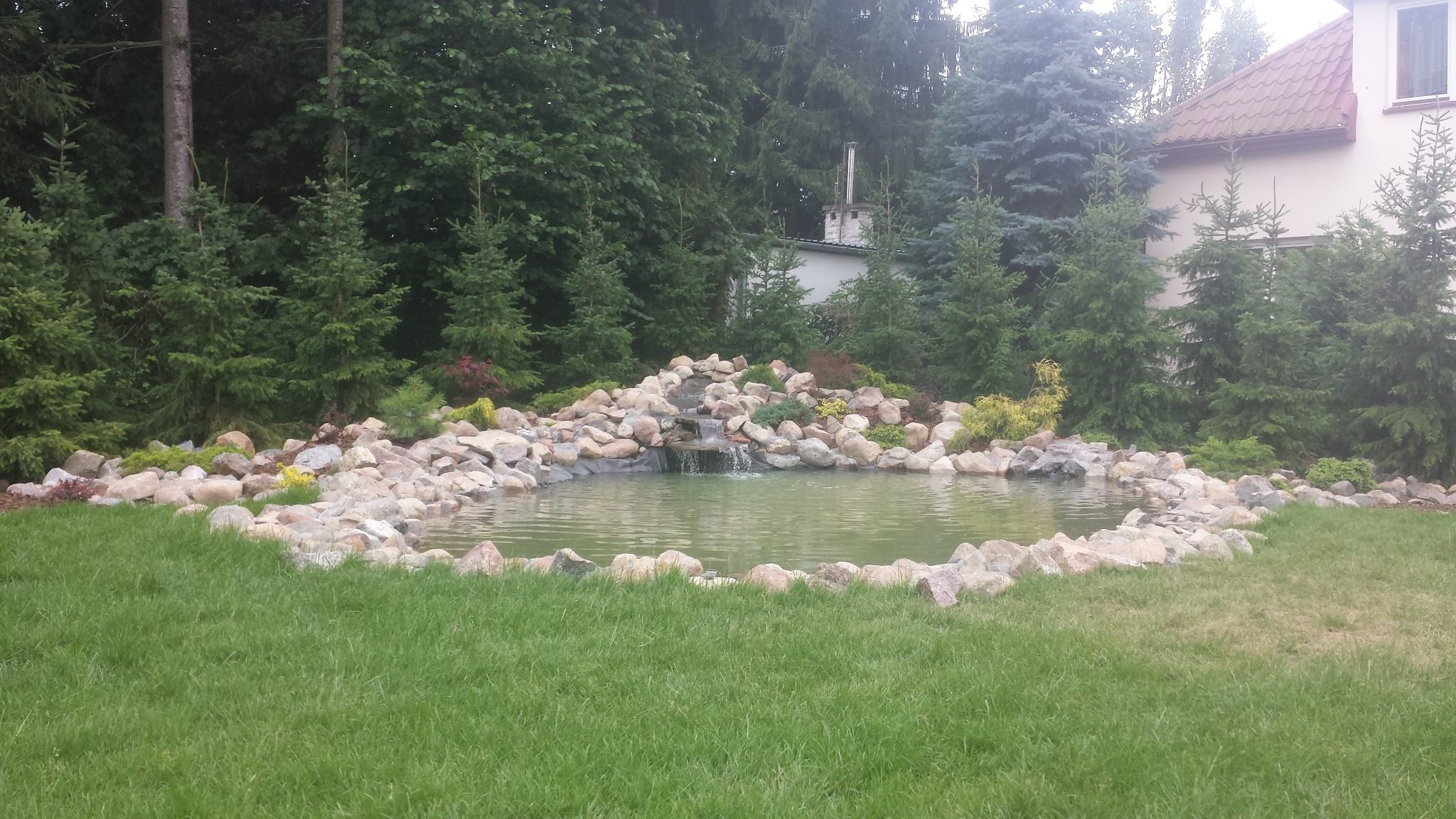 Oczko wodne z kaskadą otoczone kamieniami i zielenią na tle drzew i budynku.