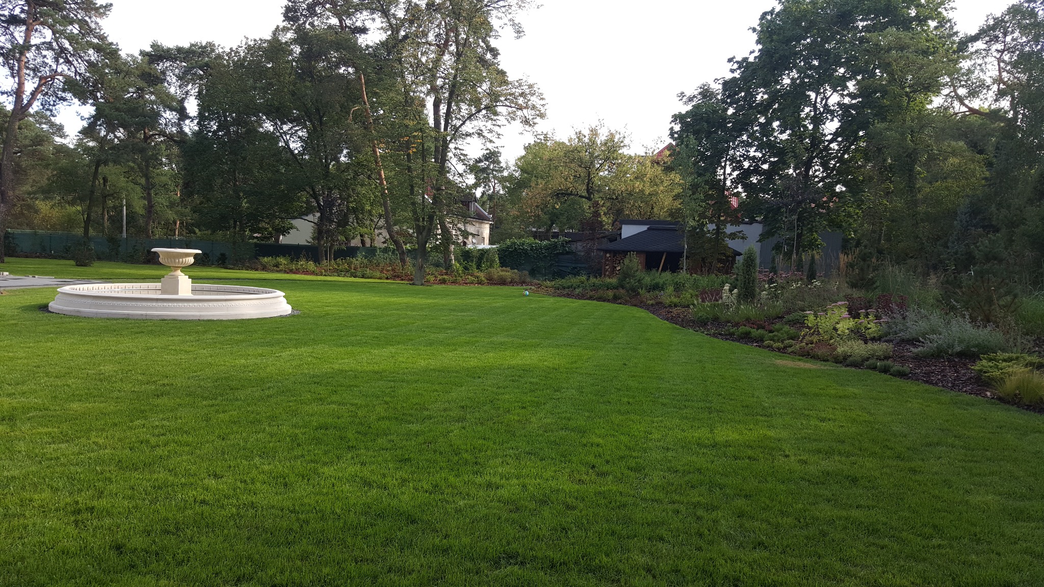 Rozległy, intensywnie zielony trawnik z fontanną w stylu klasycznym, otoczony drzewami i rabatami kwiatowymi z korą, w tle budynek.