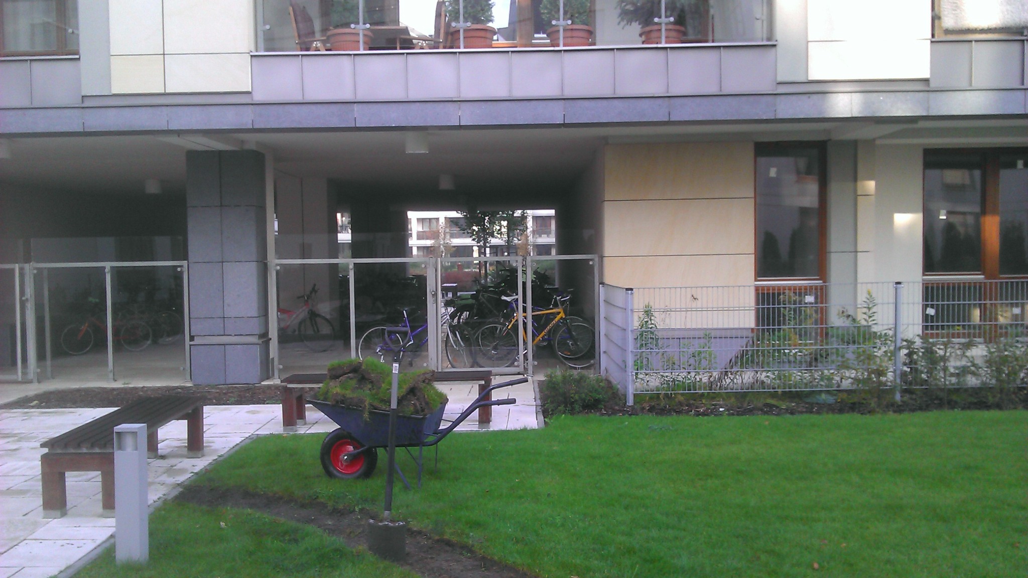 Taczka wypełniona darnią na tle nowoczesnego budynku mieszkalnego z ogrodzonym parkingiem dla rowerów, świeżo ułożona trawa widoczna w tle.