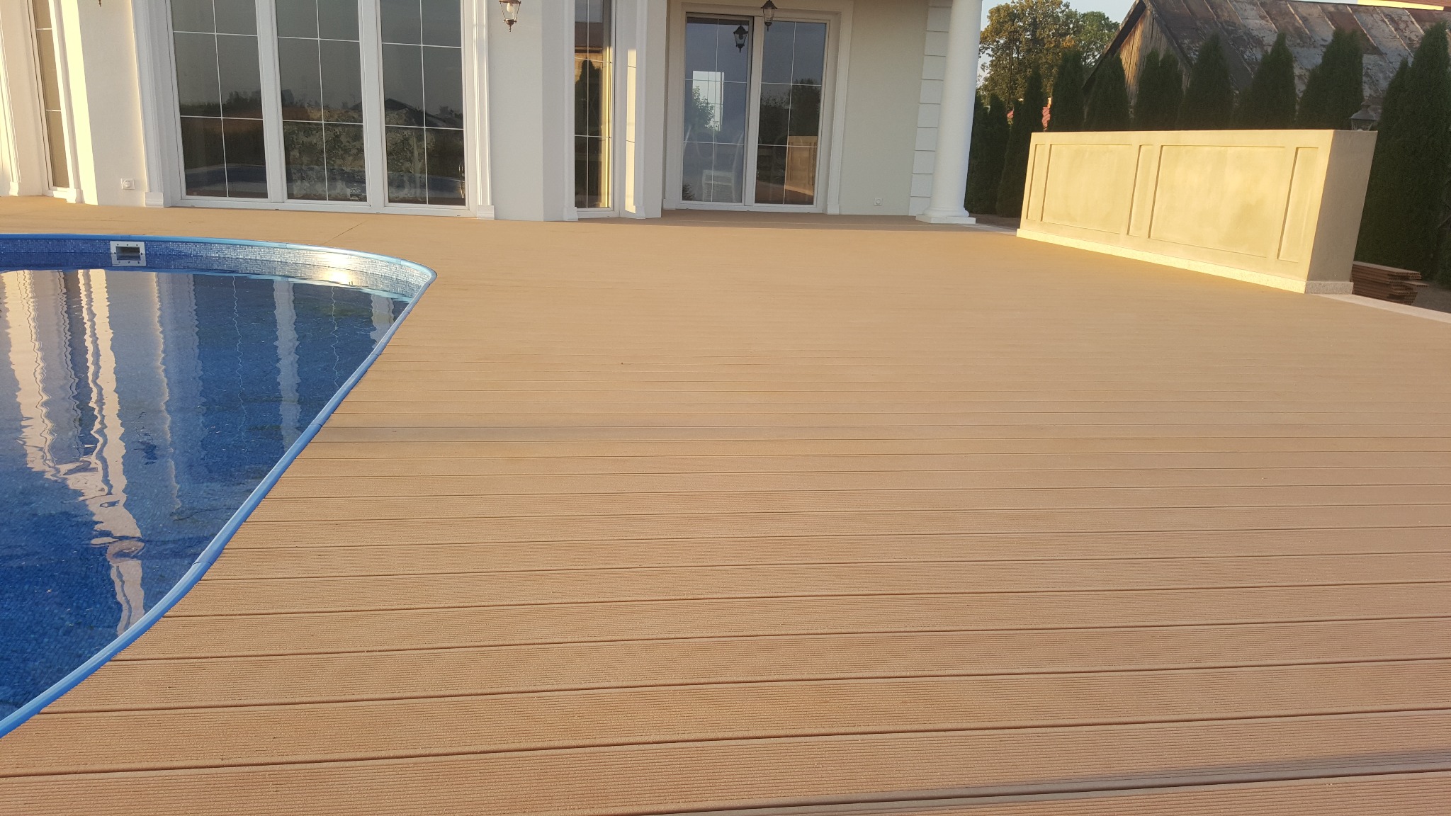 Beżowy taras kompozytowy przylegający do basenu z błękitną mozaiką, na tle białej elewacji domu z dużymi oknami.