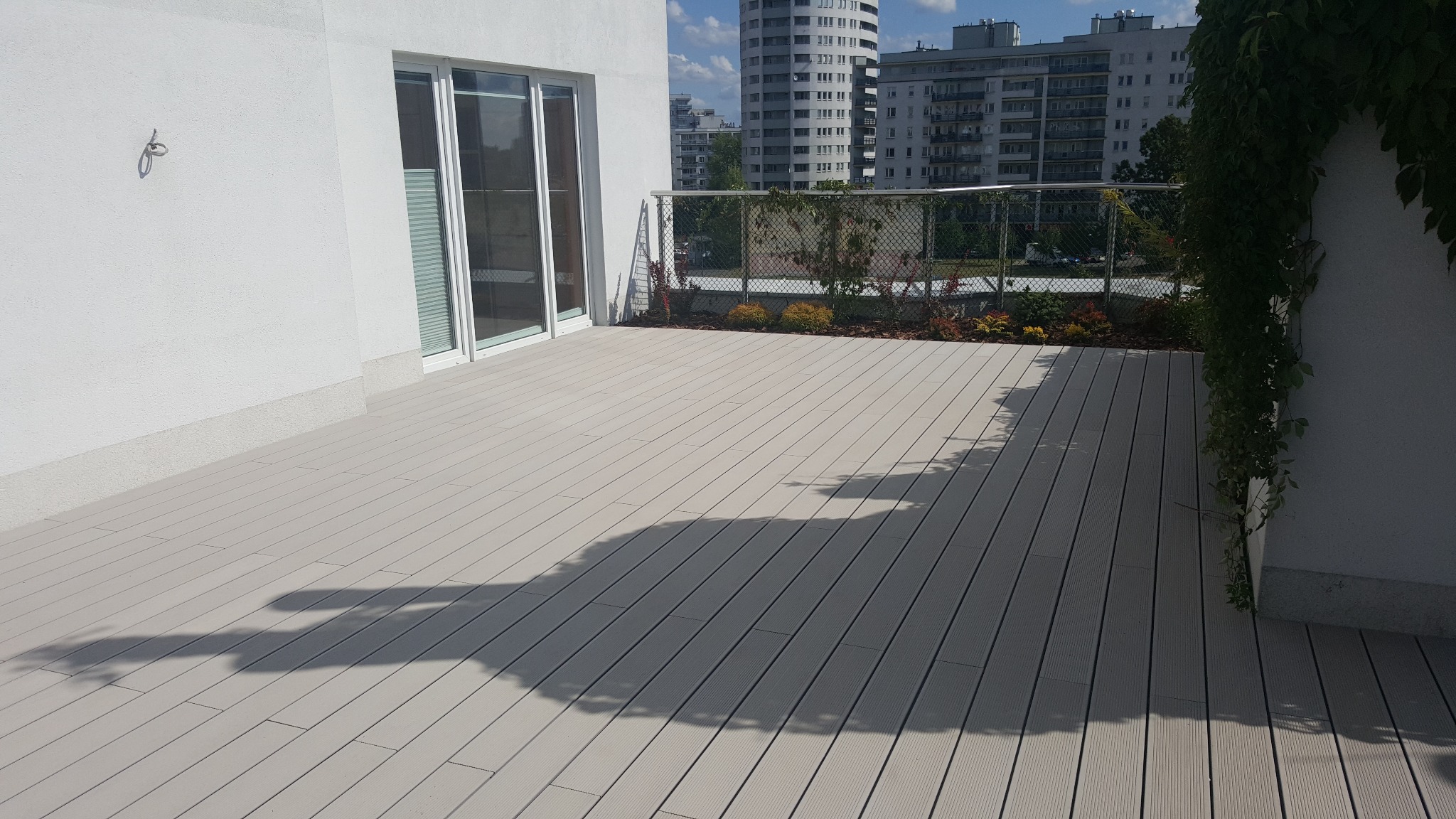 Nowoczesny taras na dachu z kompozytowych desek w odcieniach szarości, z widokiem na miejską zabudowę, roślinami ozdobnymi wzdłuż balustrady i pnączem na ścianie budynku.
