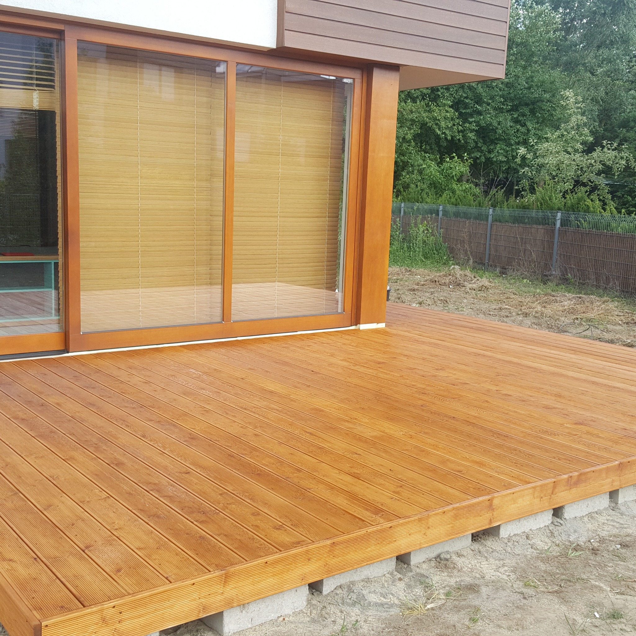 Nowo wybudowany, drewniany taras z desek kompozytowych w kolorze jasnego drewna, przylegający do budynku z przesuwnymi drzwiami tarasowymi i roletami bambusowymi, widoczny fragment trawnika...