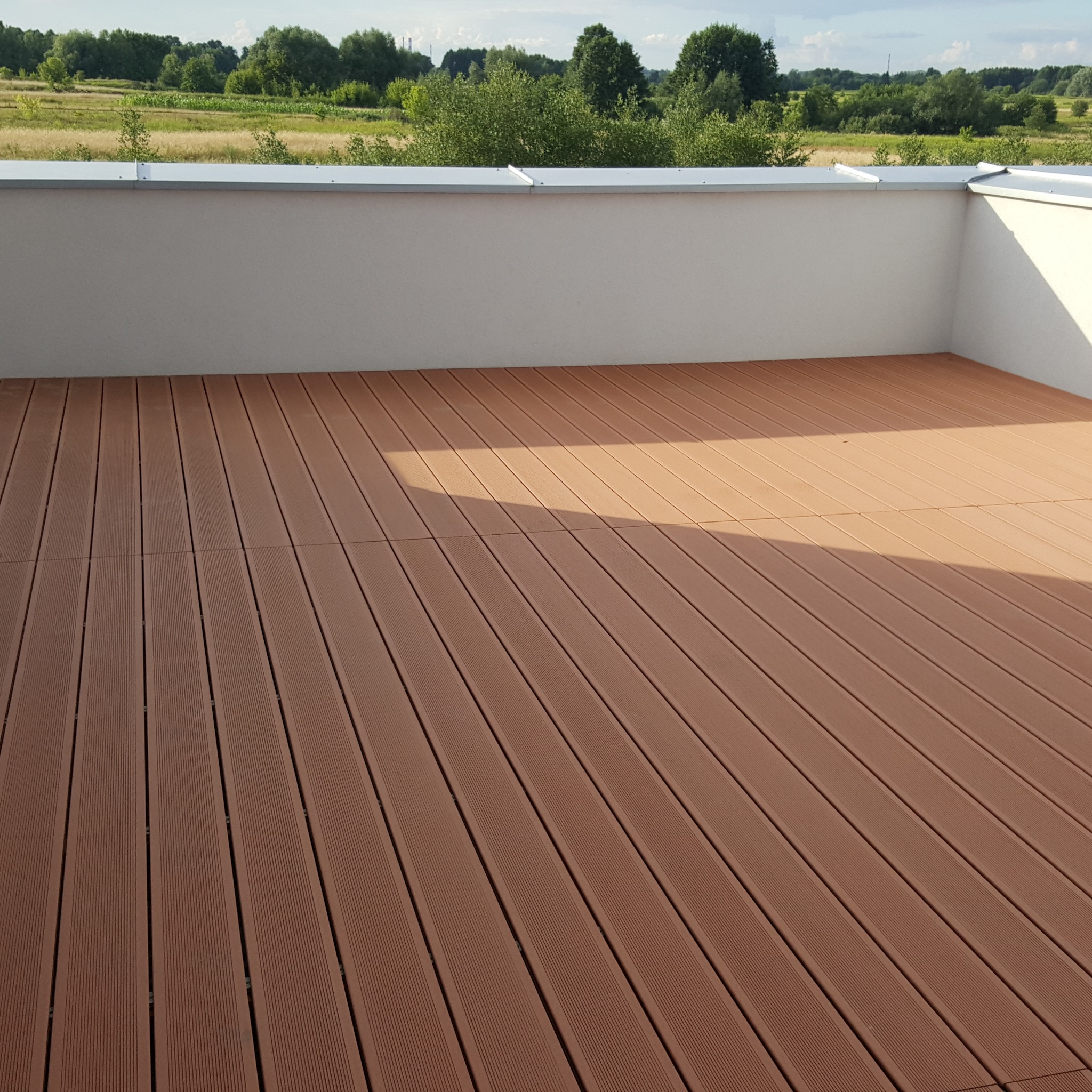 Nowoczesny taras z desek kompozytowych w odcieniach brązu, z widokiem na zielone pola i drzewa w tle.