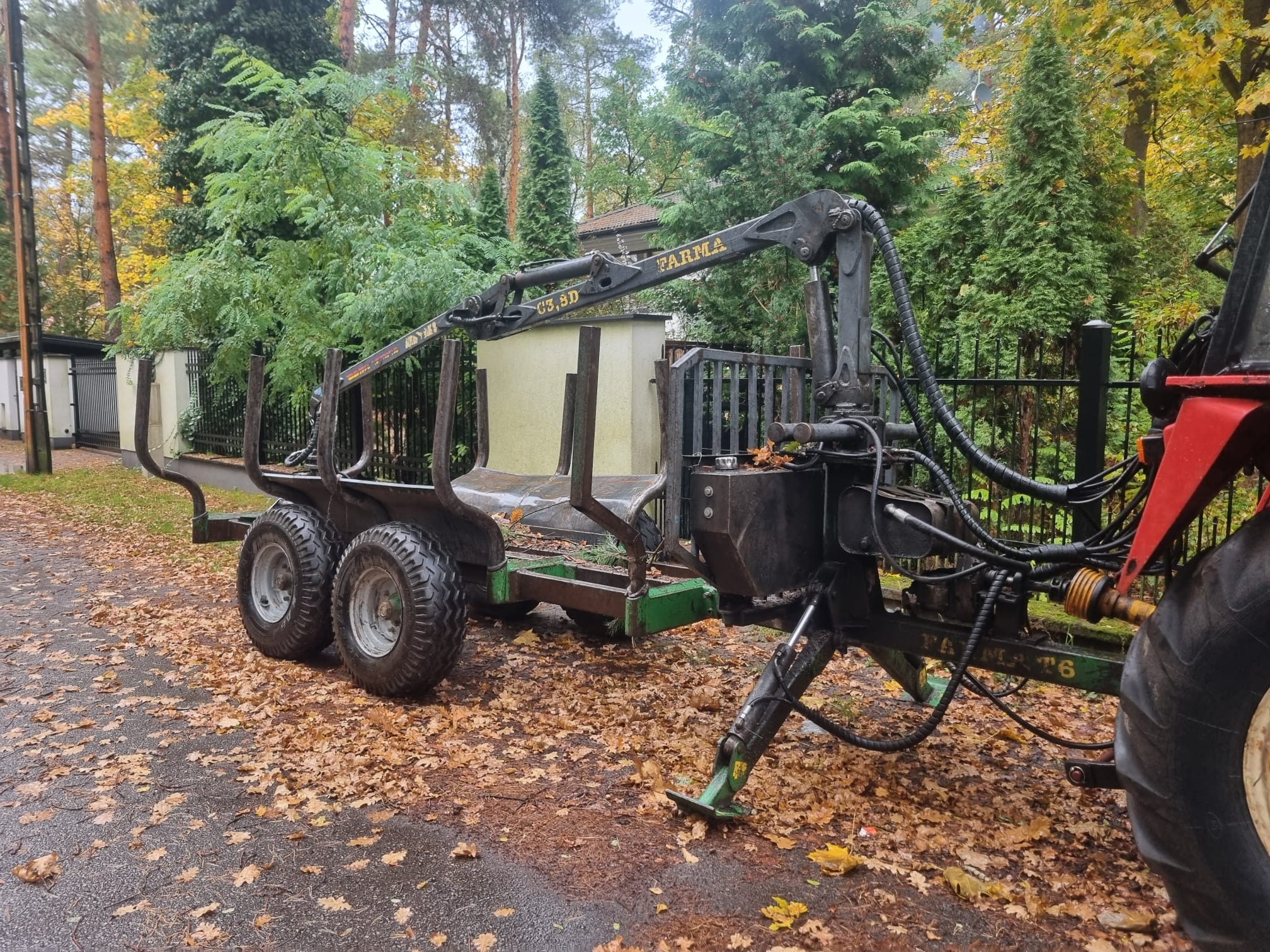 Przyczepa leśna z żurawiem marki Farma na tle jesiennego krajobrazu. Widoczne liście na ziemi i drzewa w tle. Sprzęt gotowy do pracy przy wycince lub transporcie drewna.