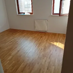 Parkiet po renowacji w pustym pokoju z dwoma oknami, widoczny grzejnik i fragment ściany