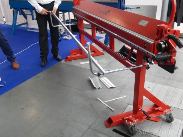 Czerwona zaginarka do blachy z oprzyrządowaniem, obsługiwana przez osobę w białej koszuli, na tle niebieskiej wykładziny podłogowej.