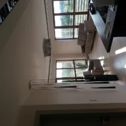 Jasne wnętrze z dużymi oknami, beżową sofą i fotelami. Na ścianie nowoczesna lampa i dekoracyjny grzejnik. Parkiet odbija światło słoneczne.