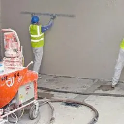 Ekipa tynkarzy w żółtych kamizelkach, nakłada tynk maszynowy na ścianę. Widoczna maszyna do tynkowania i narzędzia. Częściowo otynkowana ściana z cegieł.