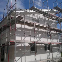 Dom jednorodzinny w trakcie tynkowania elewacji, częściowo osłonięty rusztowaniem. Widoczne fragmenty świeżego tynku i błękitne niebo.