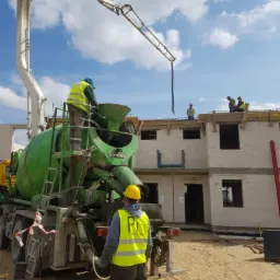 Betoniarka Stetter z wysięgnikiem pompuje beton na strop budowanego domu z białego pustaka; pracownicy w kaskach i kamizelkach odblaskowych nadzorują wylewanie betonu.