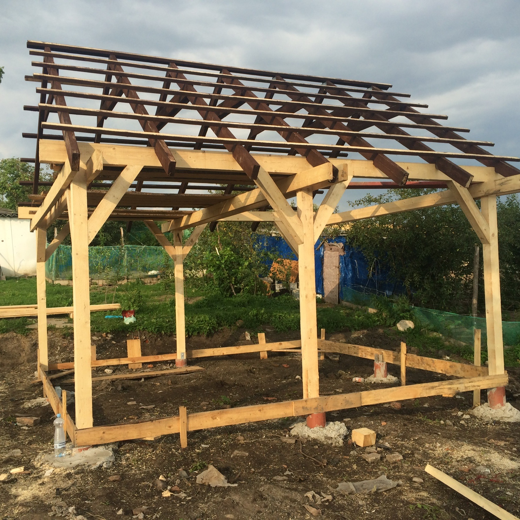 Drewniana konstrukcja altany w trakcie budowy, widoczne słupy, podwaliny i krokwie, tło z zielenią ogrodową.