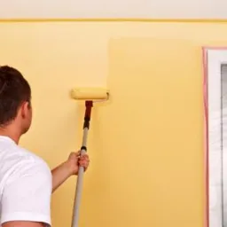 Mężczyzna w koszulce z logo 'MAXI REMONTY' maluje ścianę na żółto wałkiem, okno zabezpieczone folią.