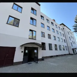 Wielopiętrowy budynek mieszkalny z jasną elewacją, brązowymi ramami okiennymi i wejściem z daszkiem, widoczny z poziomu chodnika z kostki brukowej.