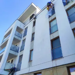 Alpiniści zabezpieczają okna folią przed malowaniem elewacji nowoczesnego budynku z balkonami, widok z dołu na błękitne niebo.