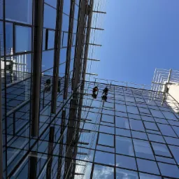 Dwóch pracowników na linach zabezpieczających, myjących szklaną fasadę nowoczesnego biurowca w słoneczny dzień, widok z dołu.